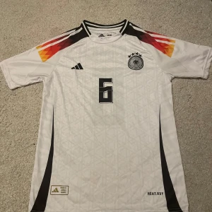 Tyskland Fotbollströja - Säljer en vit Tyskland fotbollströja med nummer 6 och Kimmich på ryggen. Tröjan har korta ärmar, svarta detaljer och mönster i rött, gult och svart på axlarna. Klassisk landslagsdesign med broderad logga och HEAT.RDY-material.