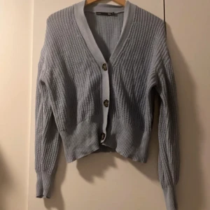 Ljusblå stickad kofta med knappar - Säljer en ljusblå stickad kofta med v-ringning och stora dekorativa knappar framtill. Koftan har lång ärm och ribbad struktur, perfekt för lager-på-lager-stil.