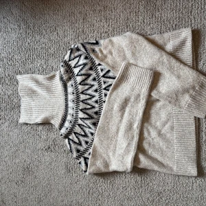 Beige stickad polotröja med mönster - Säljer en beige stickad polotröja med svart och grått zigzag-mönster över bröstet. Tröjan har lång ärm, hög polokrage och breda ribbade muddar. Perfekt för kyliga dagar!