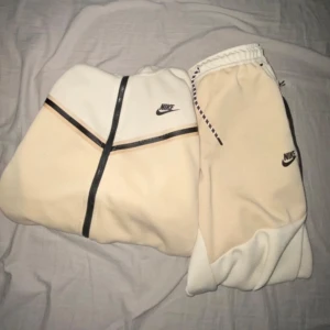 Beige och vit Nike tech - Säljer en snygg beige och vit tracksuit från Nike med huva och hel dragkedja. Jackan har svarta detaljer, broderad Nike-logga på bröstet och en praktisk ficka med dragkedja på ärmen. Perfekt för kyliga dagar.