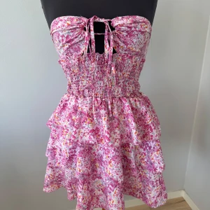 Blommig rosa volangklänning  - Supersöt kort klänning i rosa och vita toner med blommigt mönster. Klänningen har smockad midja, volanger i kjolen och knytning framtill samt halterneck. Perfekt för sommarens alla tillfällen! Storlek S och är aldrig använd☺️💕