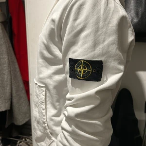 Vit hoodie från Stone Island - Säljer en stilren vit hoodie från Stone Island med deras ikoniska märke på ärmen. Tröjan har en praktisk magficka och justerbar huva med snören. Perfekt för en avslappnad stil. Nypris ca 3000kr säljer för en billig peng för att den inte används 499kr storlek är L men passar m svin bra 