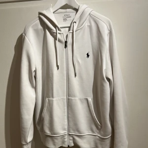 Vit hoodie från Polo Ralph Lauren - Säljer en stilren vit hoodie från Polo Ralph Lauren med dragkedja och klassisk logga på bröstet. Tröjan har en bekväm passform med långa ärmar. Använd sparsamt och är i väldigt bra skick. Finns i Linköping/Norrköping. Kom med prisförslag som passar dig!