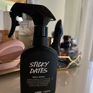 Sticky Dates Body Spray från Lush - Säljer en flaska Sticky Dates Body Spray från Lush. Den har en svart flaska med en praktisk spraypump. Doften är en sammetslen blandning av dadlar och sandelträ, perfekt för att ge en varm och inbjudande känsla. Flaskan rymmer 200 ml.