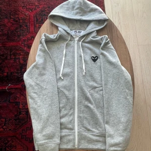 Grå CDG Kofta - Grå CDG hoodie storlek M. Svart logga. Play modellen. Nytt skick, använd två gånger. Pris kan diskuteras! Liknande kläder finns på min profil!