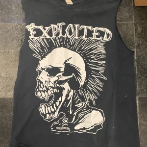 Svart ärmlös t-shirt med Exploited-tryck - Cool svart ärmlös t-shirt med stort tryck av Exploited på framsidan. Perfekt för punkälskare som vill sticka ut. T-shirten har en rå och edgy stil med ett stort skelettmotiv och texten 'Exploited' i vitt.
