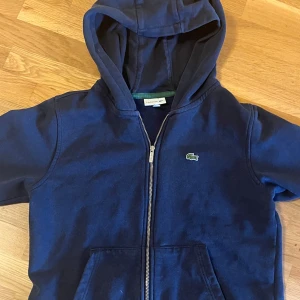 Blå hoodie från Lacoste - Säljer en snygg blå hoodie från Lacoste med dragkedja och klassisk krokodil-logga på bröstet. Perfekt för en avslappnad stil. Passar bra till både jeans och träningsbyxor.
