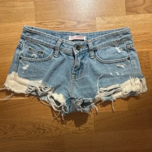 Blå jeansshorts från Blueray - Säljer ett par coola blå jeansshorts från Blueray med slitna detaljer och fransar. De har en låg midja och klassisk femficksdesign. Perfekta för en avslappnad stil!