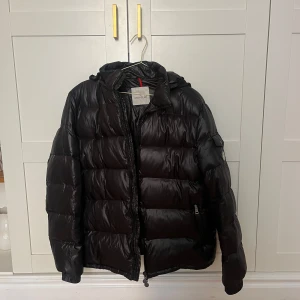 Svart Moncler dunjacka - Säljer en snygg svart Moncler dunjacka. Jackan har en glansig finish och är utrustad med dragkedja och huva. Perfekt för kyliga dagar med sin varma och bekväma design. Den har även en unik etikett med serietidningsmotiv på insidan. Tagit över efter min bror & säljer den då jag inte använder den så ofta. Min bror är strl L, jag är strl M. Passar M & L bäst beroende på fit som önskas. Herr modell men Unisex skulle jag säga.