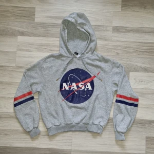 Grå NASA hoodie med tryck - Säljer en grå hoodie från Divided med stort NASA-tryck på framsidan. Tröjan har en klassisk huva och ribbade muddar. Ärmarna har röda och blå ränder som ger en sportig look. Perfekt för alla rymdentusiaster! 🚀