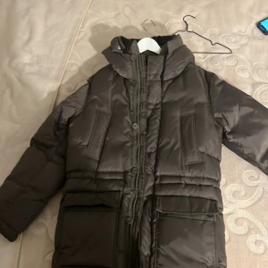 Brun dunjacka från J.Lindeberg - Säljer en brun dunjacka från J.Lindeberg. Jackan har en hög krage och en avtagbar huva. Den är långärmad med dragkedja och knappar framtill. Perfekt för kalla dagar. Riktigt bra kvalitet då den köptes i butik för 2990kr. Mitt pris 2500kr, är ej hugget i sten!…….Mvh Emilia!