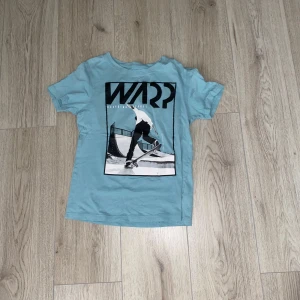 Blå t-shirt  - Säljer en cool blå t-shirt från Warp med ett stort tryck av en skateboardåkare på framsidan. Perfekt för skate-entusiaster! T-shirten är kortärmad och gjord i ett mjukt material.