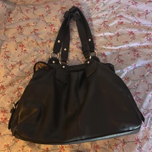 Svart axelväska med guld detaljer  - Snygg svart axelväska i syntetmaterial med guld detaljer. Väskan har flera praktiska fack. Rätt så stor i storleken, får platts med mycket 