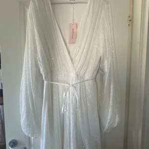 Vit glittrig klänning - Säljer en elegant vit klänning med glittrande paljetter och v-ringad design. Klänningen har långa ärmar och ett knytband i midjan för en smickrande passform. Perfekt för speciella tillfällen! Jag beställde den förra sommaren när jag var gravid och skulle gifta mig men bestämde mig för en annan klänning. Denna är aldrig använd och lappen är kvar.