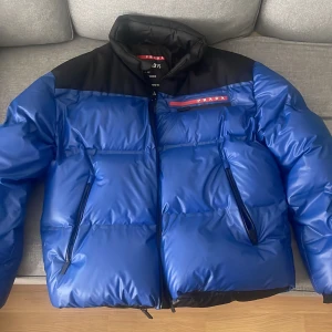 Blå och svart pufferjacka från Prada - Snygg blå och svart linea rosa pufferjacka från Prada med dragkedja och två fickor framtill. Jackan har en hög krage och är perfekt för kyligare väder. Den har en stilren design med Pradas logotyp på bröstet. Varan är original (äkta) och köpt på Prada Stockholm på Birger Jarlsgatan  nypris 34900kr