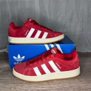 Röda Adidas Campus sneakers - Snygga röda Adidas Campus sneakers med klassiska vita ränder och snörning. Skorna har en stilren design med en bekväm sula och är perfekta för en sportig look. De är tillverkade i mocka och har en vit yttersula som ger en fin kontrast.