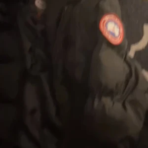 Svart dunjacka från Canada Goose - Säljer en svart dunjacka från Canada Goose med deras ikoniska märke på ärmen. Jackan har en pälskantad huva och är perfekt för kalla vinterdagar. Den är långärmad och har en dragkedja framtill.