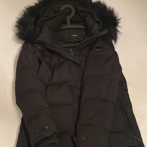Svart dunjacka med fuskpäls - Snygg svart vinterjacka från ROCKANDBLUE med avtagbar fuskpäls på huvan. Jackan har dragkedja och och fickor.Perfekt för kalla vinterdagar.