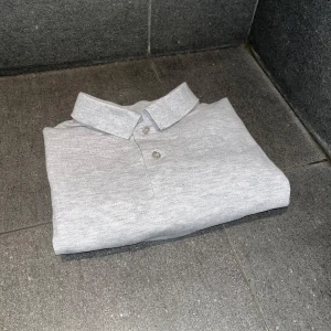 Grå pikétröja från Dressmann - Säljer en stilren grå pikétröja från Dressmann. Tröjan har en klassisk krage med knappar och är kortärmad. Perfekt för en avslappnad look. Fotomodellen är 174