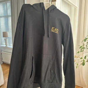 Svart hoodie från Emporio Armani - Snygg svart hoodie från Emporio Armani med gul logga på bröstet. Tröjan har en klassisk design med huva och känguruficka framtill. Perfekt för en stilren och bekväm look.