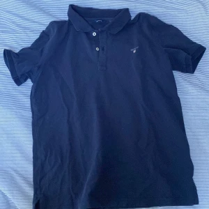 Blå pikétröja från GANT - Säljer en stilren svart pikétröja från GANT med klassisk krage och knappar framtill. Tröjan har en liten broderad logotyp på bröstet. Perfekt för en avslappnad men ändå snygg look.