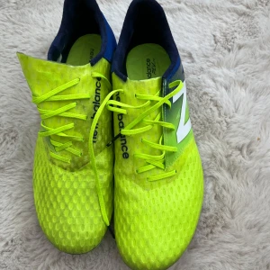 Neongula fotbollsskor från New Balance - Snygga neongula fotbollsskor från New Balance med blå detaljer och snörning. Skorna har en mönstrad ovansida och robusta dobbar för bra grepp på planen. Perfekta för den som vill sticka ut på fotbollsplanen!