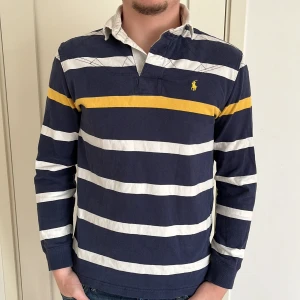 Randig pikétröja från Polo Ralph Lauren - Säljer en snygg randig pikétröja från Polo Ralph Lauren i marinblått och vitt med en gul rand över bröstet. Tröjan har långa ärmar och en klassisk krage. Perfekt för en stilren look.