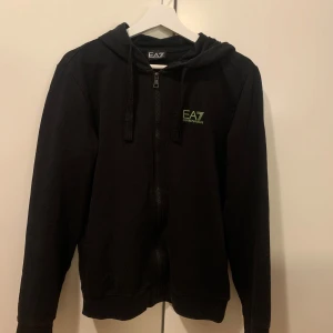 Svart hoodie från Emporio Armani - Säljer svart zip up hoodie från Emporio Armani med dragkedja och huva. Den har en liten grön logga på bröstet och snören vid huvan. Den har ett super litet hål vid ena handleden, cirka 1 cm stort. Men det är inget man riktigt märker.💕