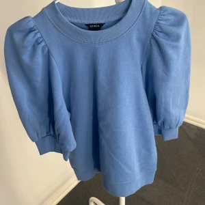 Blå tröja från Lindex - Säljer en söt blå tröja från Lindex med puffiga ärmar och rund halsringning. Perfekt för en stilren och bekväm look. Tröjan har ribbade muddar vid ärmslut och nederkant. Helt ny så inte använd!