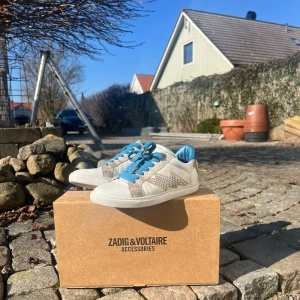 Vita sneakers med blå detaljer från Zadig & Voltaire - Snygga vita sneakers med blå snörning och häl från Zadig & Voltaire. Skorna har en cool blixtdetalj i guld och silver på sidan. Perfekta för att ge din outfit en extra edge.