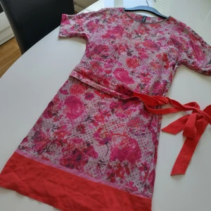 Blommig klänning från Marc Cain - Säljer en färgglad blommig klänning från Marc Cain i SIDEN. Klänningen har korta ärmar och ett vackert blommönster i rosa och röda nyanser med prickar. Den har en röd kant nedtill och ett matchande knytband i midjan.