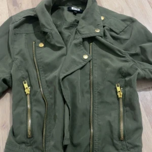 Grön jacka från Bik Bok - Snygg grön jacka från Bik Bok med dragkedjor och knappar i guld. Helt slutsåld och är knappt använd. Passar perfekt nu i våren. Skriv om fler bilde
