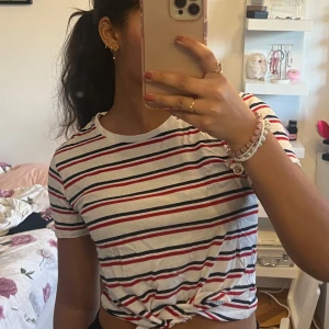 Randig t-shirt från H&M - Säljer en vit t-shirt med röda och svarta ränder från H&M. Den har korta ärmar och en knutdetalj framtill som ger en trendig look. Perfekt för en avslappnad stil.