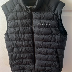 Svart dunväst från Sail Racing - Snygg svart dunväst från Sail Racing med dragkedja framtill. Västen har en stilren design med logotyp på bröstet och ryggen. Perfekt för kyligare dagar när du vill hålla värmen utan att kompromissa med stilen.