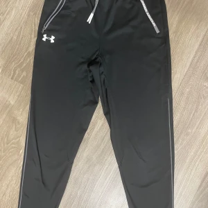 Under armour byxor  - Säljer ett par svarta trendiga träningsbyxor från Under Armour med dragsko i midjan och dragkedjefickor. Byxorna har en lös passform och är perfekta för sportiga aktiviteter. Under Armour-loggan syns på ena benet.