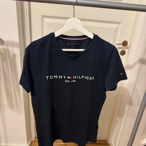 Svart t-shirt från Tommy Hilfiger - Snygg s t-shirt från Tommy Hilfiger med tryck på framsidan. Perfekt för en avslappnad stil. Klassisk rund hals och korta ärmar. Storlek: S Nypris: 500kr