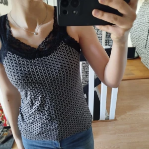 Svart och vit linne med spetsdetaljer - Snyggt svart och vitt linne med spetsdetaljer vid halsringningen. Mönstrat med små cirklar och har en tight passform. Perfekt för en stilren look.