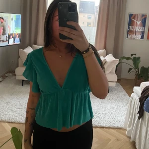 Grön v-ringad blus - Säljer en snygg grön blus med v-ringning och volangdetaljer. Toppen är croppad och har korta ärmar, perfekt för en somrig look. Passar bra till både jeans och kjol.