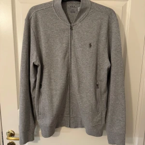 Grå tröja från Polo Ralph Lauren - Säljer en stilren grå tröja från Polo Ralph Lauren med dragkedja framtill. Tröjan har långa ärmar och en liten broderad logga på bröstet. Perfekt för en avslappnad look.