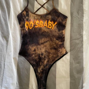 Brun ribbad body med text - Trendig brun body med ribbad textur och texten '90's BABY' i eldigt gult och rött. Den har smala axelband som korsas i ryggen för en stilren look. Perfekt för en retro vibe på stranden!