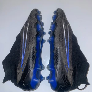 Svarta och blå fotbollsskor från Nike - Snygga fotbollsskor från Nike i svart och blått med en strömlinjeformad design. Skorna har en stickad överdel för optimal passform och en robust sula med dobbar för bra grepp på planen. Perfekta för fotbollsträning och matcher.