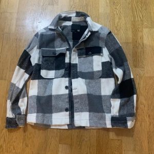 Rutig overshirt från 157 - Snygg rutig overshirt från 157 i svart, grått och vitt. Jackan har knappar framtill och två bröstfickor med lock. Perfekt för lager-på-lager-stil under höst och vår.