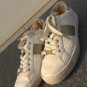 Vita sneakers från Michael Kors - Snygga vita sneakers från Michael Kors med gulddetaljer och snörning. Skorna har en quiltad textur och en bred guldfärgad rand på sidan. Perfekta för en stilren look.