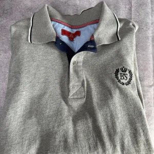 Grå pikétröja från BS - Säljer en stilren grå pikétröja från BS med broderad logga på bröstet. Tröjan har långa ärmar och en klassisk krage med kontrastfärger i blått och rött. Perfekt för en avslappnad men ändå uppklädd look.