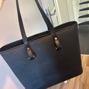 Svart axelväska med dragkedja - Snygg svart axelväska i syntetmaterial med praktisk dragkedja. Perfekt för att bära med sig allt man behöver. Stilren design med långa handtag för enkel bärning.