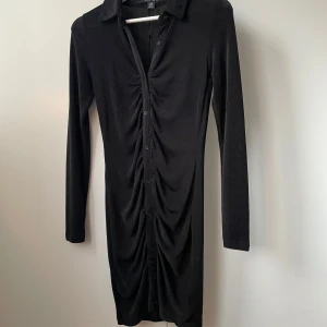 Svart långärmad klänning med knappar - Elegant svart klänning från New yorker med långa ärmar och knappar framtill. Klänningen har en stilren design med rynkdetaljer som ger en smickrande passform. Perfekt för en stilfull look.