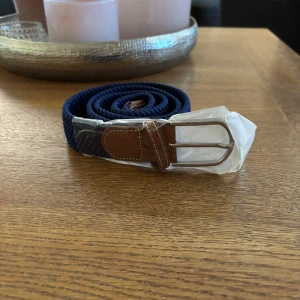 Blått flätat bälte med brunt läder - Snyggt blått flätat bälte med detaljer i brunt läder och ett silverfärgat spänne. Perfekt för att ge en stilren touch till din outfit. Bältet är flexibelt och passar de flesta storlekar.