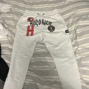 Vita mjukisbyxor från Hoodrich - Säljer ett par vita mjukisbyxor från Hoodrich med coola broderade detaljer och logotyper. Byxorna har snörning i midjan och en avslappnad passform. Perfekta för en chill dag hemma eller ute.