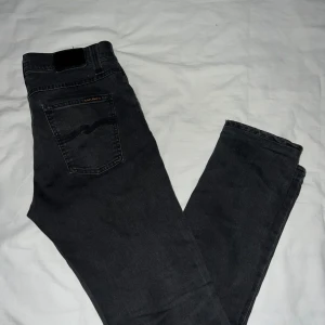 Svarta jeans från Nudie Jeans - Snygga svarta jeans från Nudie Jeans med klassisk femficksdesign och knappgylf. Jeansen har en rak passform och är tillverkade i slitstarkt denim. Perfekta för en stilren look.