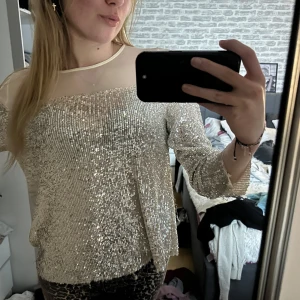 Glittrig offshoulder-top - Säljer en glittrig offshoulder-top i silver med långa ärmar. Perfekt för en festlig kväll! Toppen har en transparent detalj vid halsen som ger en extra touch av elegans.
